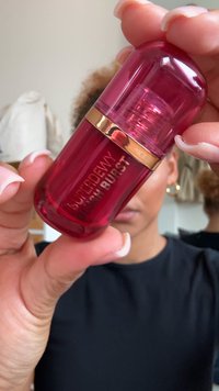 Ljusröd cylindrisk flaska med ett guld band, märkt "SUPERDEWY BLUSH BURST", med en vridkork och en slät, glänsande yta.