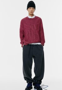 Giovane che indossa un berretto nero, un maglione oversize bordeaux con lettering in rilievo, pantaloni larghi grigi scuri e stivali neri con lacci blu.