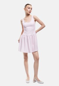 Una mujer está de pie, vistiendo un vestido de cuadros liviano color rosa claro con un dobladillo con volantes y zapatos de tacón bajo blancos, mirando de lado.