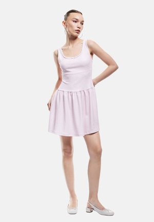 Eine Frau steht da und trägt ein ärmelloses, hellrosa kariertes Kleid mit einem Rüschenabschluss und weißen, flachen Schuhen, während sie zur Seite schaut.