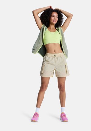 Femme souriante aux cheveux bouclés portant un soutien-gorge de sport vert clair, un short cargo beige, un gilet polaire gris, des chaussettes blanches et des baskets roses, bras levés.