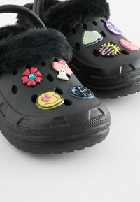 Sabots noirs avec une bordure en fausse fourrure, ornés de charms en caoutchouc colorés, comprenant des fleurs et des motifs ludiques, ainsi que des trous de ventilation.