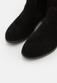 Par de botines de ante negro con puntas redondeadas y suelas de goma negra sobre un fondo blanco.
