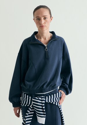 Jeune femme portant un pull zippé bleu marine et un pull rayé noué autour de la taille, debout devant un fond clair uni.