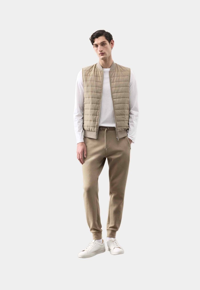 Chaleco acolchado beige sobre una camisa blanca, combinado con pantalones jogger beige y zapatillas blancas. Textura suave, diseño ajustado y acentos sutiles.