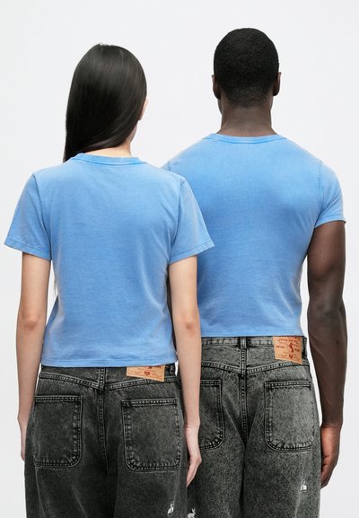 Ανοιχτό μπλε βαμβακερά T-shirt με κοντά μανίκια, κομμένα στο ύψος της μέσης. Φοριούνται με ψηλόμεσα, σκουρόχρωμα τζιν από τζιν ύφασμα με ορατές ετικέτες μάρκας.