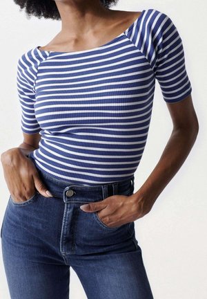 Femme portant un top ajusté à rayures horizontales bleues et blanches et un jean taille haute bleu foncé, les mains dans les poches.