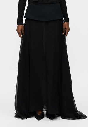 Žena nosi dlhú, splývavú čiernu sukňu s vrstvenou látkou a čierne lodičky s špicatou špičkou, stojaca pred jednoduchým svetlým pozadím.