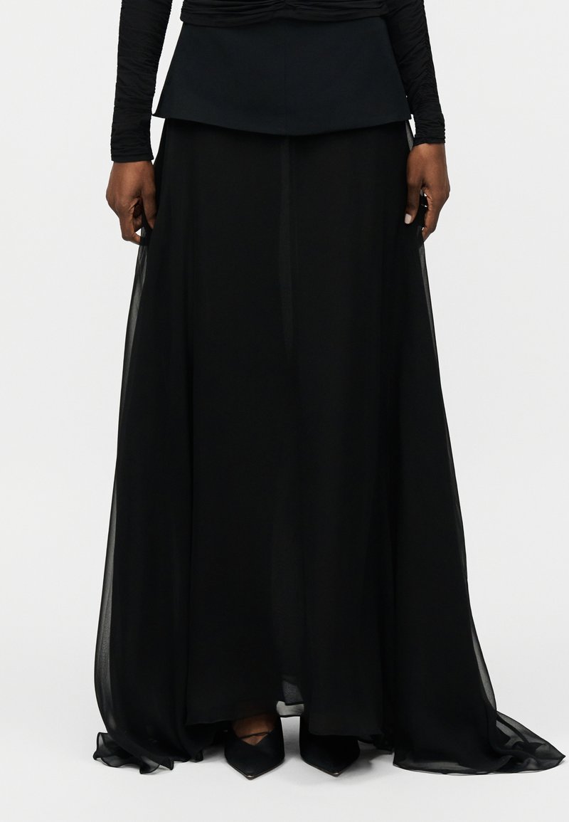 Kvinde iført en lang, flydende sort nederdel med lagdelt stof og sorte spidse hæle, stående mod en ensfarvet lys baggrund.