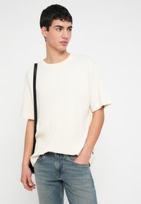 Νεαρός άνδρας με κοντά σκουρόχρωμα μαλλιά φοράει μπλουζάκι με ριμπ ύφανση σε κρεμ χρώμα και κοντά μανίκια, μπλε τζιν και μαύρο σακίδιο ώμου, κοιτάζοντας πλάγια.