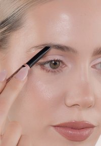 Ein Augenbrauenstift wird mit einer schlanken schwarzen Spitze auf natürlichen Augenbrauen aufgetragen, Nahaufnahme eines Gesichts mit heller Haut und zartem rosa Lippenstift.