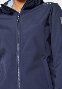 Veste softshell bleu marine avec une fermeture éclair à l'avant, une capuche et deux poches latérales. Présente une texture lisse et mate ainsi que des accents de logo sur l'épaule.