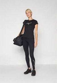 Juoda sportinė marškinėliai su sidabro spalvos logotipu, derinami su juodais tampriais, kuriuose yra sidabro akcentas. Modelis laiko juodą sportinį krepšį.