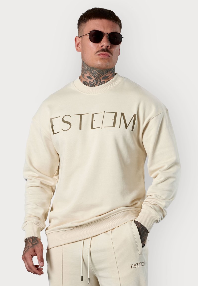 Sweatshirt beige avec un col rond, arborant un grand logo marron "ESTEEM" sur la poitrine, des poignets côtelés et une coupe décontractée.