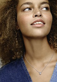 Srebrna ogrlica s pendantom v obliki srca, ki vsebuje majhen diamantni akcent, nošena na modelu s kodrastimi lasmi na ozadju iz džinsa.