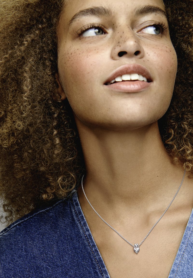 Srebrna ogrlica s pendantom v obliki srca, ki vsebuje majhen diamantni akcent, nošena na modelu s kodrastimi lasmi na ozadju iz džinsa.