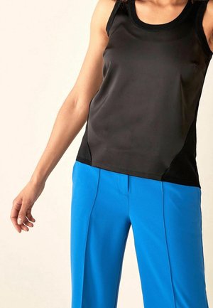 Kvinde iført en sort ærmeløs top og klare blå bukser, stående med en arm afslappet langs siden mod en ensfarvet baggrund.