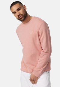 Sudadera de color rosa claro con cuello redondo y puños de canalé, que presenta un pequeño logo blanco en el pecho, fabricada con material suave.