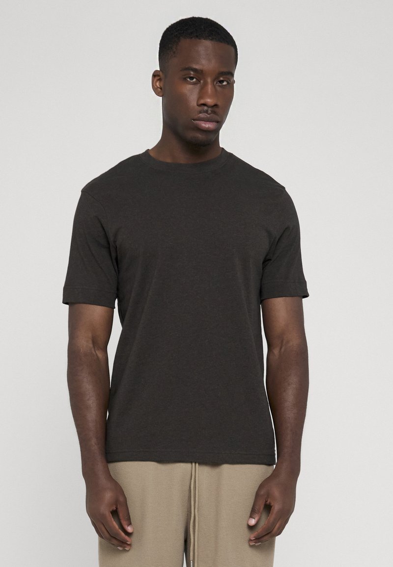 Homme debout, face à la caméra, portant un t-shirt gris foncé uni à manches courtes et un pantalon beige texturé sur un fond gris clair.