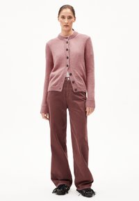 Eine Frau trägt eine hellrosa Knopfpulloverjacke, kastanienrote weite Hose und schwarze Fransen-Chic-Loafer und steht vor einem schlichten weißen Hintergrund.