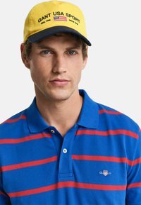 Sininen pikeepaita, jossa on punaisia vaakaraitoja, sekä sininen ja keltainen lippalakki, jossa lukee "GANT USA SPORT" ja joka sisältää Amerikan lipun emblem.
