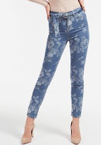 Jeans de mezclilla con estampado floral en azul claro, que presentan un diseño ajustado, detalle de cinturón y una textura suave, combinados con tacones nude.