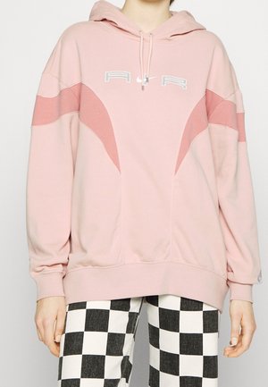 Personne portant un sweat à capuche rose ample avec le texte "AIR" et des panneaux roses plus foncés incurvés, associé à un pantalon à carreaux noir et blanc.