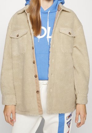 Osoba ubrana w beżową, zapinaną na guziki polarową kurtkę na niebieską bluzę z kapturem białymi sznurkami i białe spodnie z kolorowymi naszywkami.