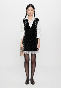 Μαύρο βελούδινο φόρεμα με λευκές βούλες και φούντες στο τελείωμα, συνδυασμένο με λευκή πουκαμίσα με γιακά, μαύρες καλσόν και καφέ φλατ παπούτσια. Κρατάει μια μικρή τσάντα.