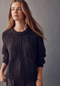 Brun, tekstureret striktrøje med rund halsudskæring og ribstrikkede manchetter, der har et diskret geometrisk mønster. Modellen har et ur på.