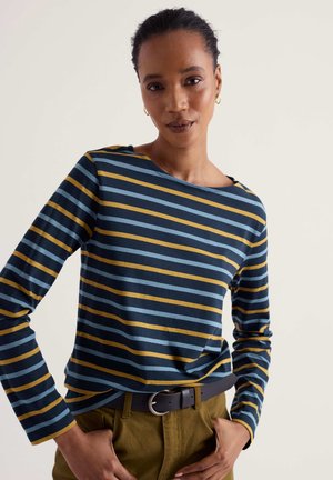 Frau mit zurückgebundenen Haaren trägt ein langärmliges Shirt mit navy-, blauen und senffarbenen Streifen sowie eine olivfarbene Hose, Hände in den Taschen, neutraler Hintergrund.