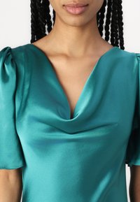 Top in satin teal con maniche corte a palloncino, con una scollatura a V e un'elegante caduta sul petto. Texture liscia e lucida in tutto il capo.