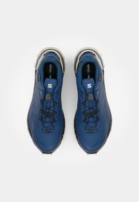 Des chaussures de sport bleues avec une tige synthétique texturée, des lacets noirs et une semelle grise rembourrée. Présente un petit logo sur le côté et un matériau imperméable.