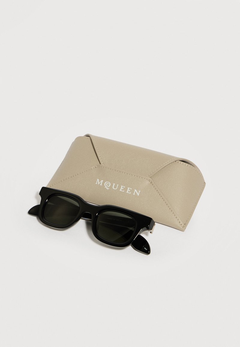 Occhiali da sole rettangolari neri appoggiati su una custodia in pelle beige con il marchio McQueen, su sfondo bianco.
