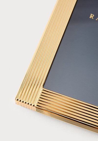 Arany színű fém képkeret bordázott mintázattal, sík szegéllyel és sötét üveg középponttal. Tiszta vonalak és tükröződő felület részletezése.