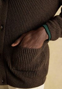 Barna bordázott kardigán gombos záródással és zöld akcentussal a csuklón; bal oldalon látható egy oldalsó zseb. Puha tapintású.