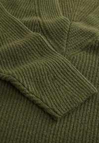 Jersey de punto verde oliva con textura acanalada y detalles de costuras diagonales. La tela parece suave y cálida, con una sensación acogedora.