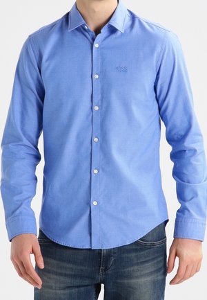 Mężczyzna ubrany w jasnoniebieską, zapinaną na guziki, koszulę z długim rękawem i małym logo „BOSS” na piersi oraz ciemnoniebieskie dżinsy.