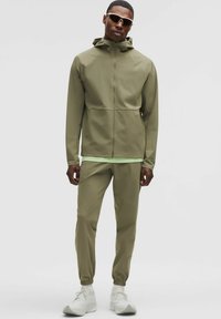 Olivgrüne Zip-Jacke mit Kapuze, kombiniert mit passenden, schmal geschnittenen Hosen. Das Material wirkt leicht mit einer glatten Textur. Weiße Sneakers.