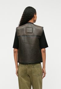 Bruine leren vest met een gestructureerde afwerking, voorzien van een label aan de achterkant, zwarte bies en gestyled met een zwart T-shirt met korte mouwen en olijfgroene broek.
