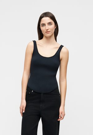 Tânără cu păr lung și întunecat, purtând un top negru fără mâneci, cu model nervurat, și pantaloni negri, stând pe fundal simplu, deschis la culoare.