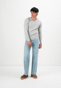Férfi világosszürke V nyakú pulóverben, világoskék farmernadrágban és barna slip-on cipőben áll a fehér háttér előtt.