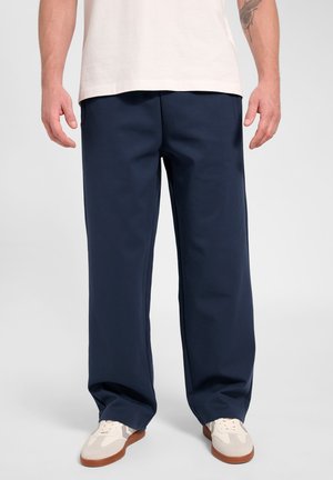 Person iført marineblå vidde bukser, hvide sneakers med brune såler og en hvid skjorte, stående mod en ensfarvet baggrund.