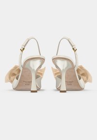 Λευκά ψηλοτάκουνα σανδάλια με λαμπερή μεταλλική τακούνι και μπεζ �υφασμάτινες λεπτομέρειες στο πίσω μέρος. Ρυθμιζόμενο λουράκι στον αστράγαλο με χρυσό κούμπωμα.