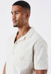 Camisa de botón de manga corta en beige claro con un patrón texturizado. Presenta un bolsillo en el pecho con un branding sutil y un cuello relajado.