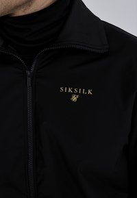 Čierna bunda vyrobená z hladkého materiálu s vysokým golierom, s zlatým logom "SIKSILK" a zipsom na zapínanie.