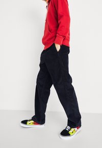 Personne portant un sweat à capuche rouge, un pantalon en velours côtelé bleu foncé et des baskets noires avec des motifs de flammes jaunes et rouges, debout les mains dans les poches.