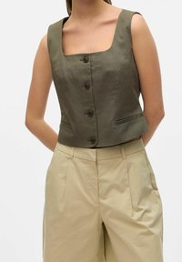 Oliwkowy skrócony top z kwadratowym dekoltem, pięcioma brązowymi guzikami i kieszeniami po bokach, zestawiony z beżowymi spodniami o szerokich nogawkach.