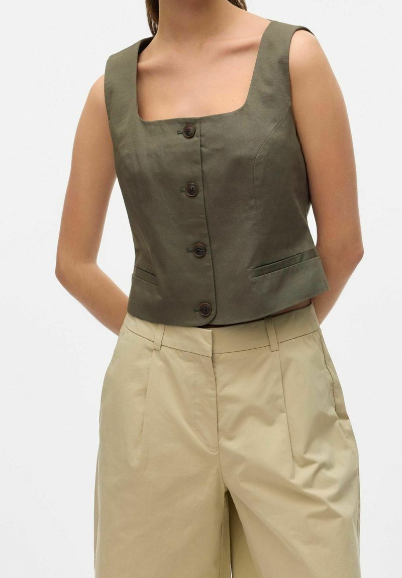 Oliwkowy skrócony top z kwadratowym dekoltem, pięcioma brązowymi guzikami i kieszeniami po bokach, zestawiony z beżowymi spodniami o szerokich nogawkach.