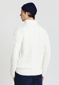 Weißer, gestrickter Pullover mit hohem Kragen und gerippten Bündchen, kombiniert mit einer marineblauen Mütze. Die Strickstruktur ist prominent und detailliert.